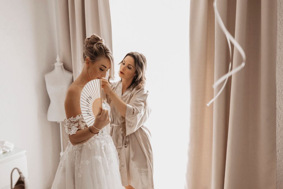 Hochzeitsfotografie - Braut wird beim Ankleiden von einer Stylistin in einem hellen Raum unterstützt.