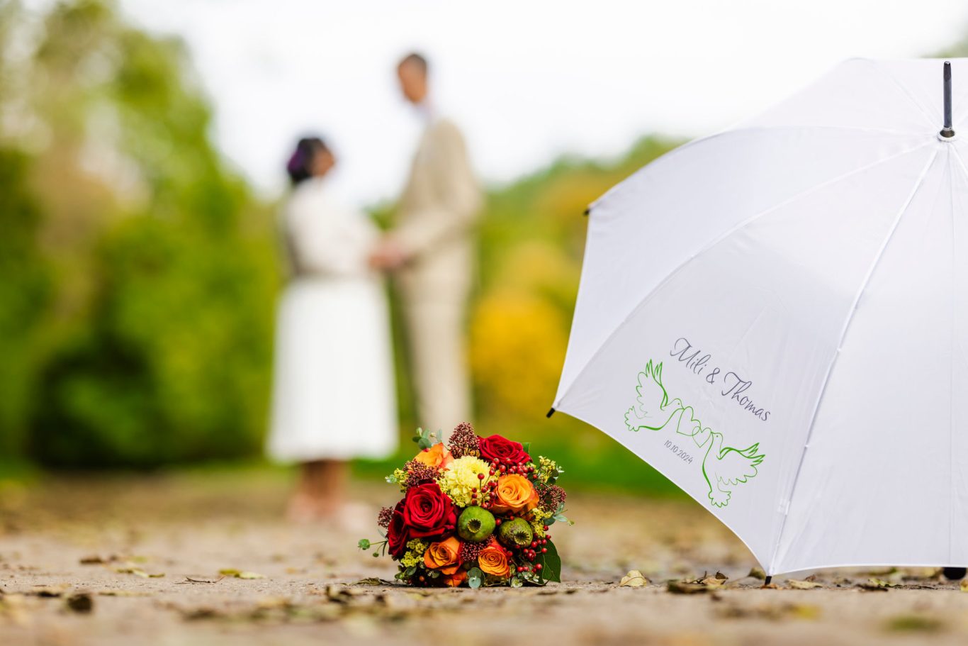 Brautpaar steht zusammen im Freien, mit einem Regenschirm und einem Blumenstrauß im Vordergrund.